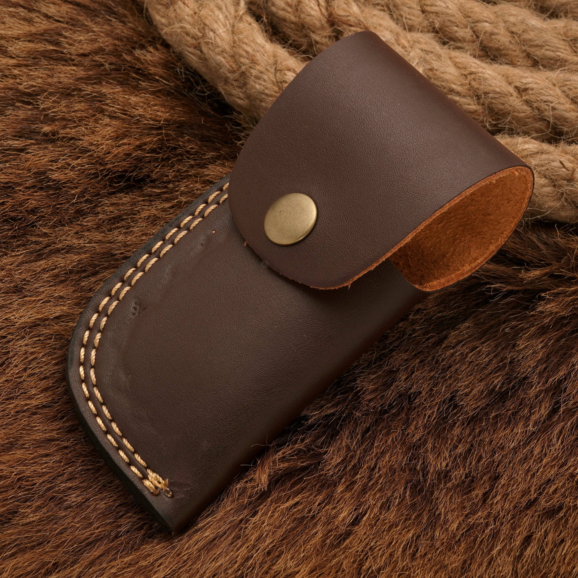 Brown leather sheath with brass button closure, designed to securely hold a Damascus pocket knife, displayed on a fur background.